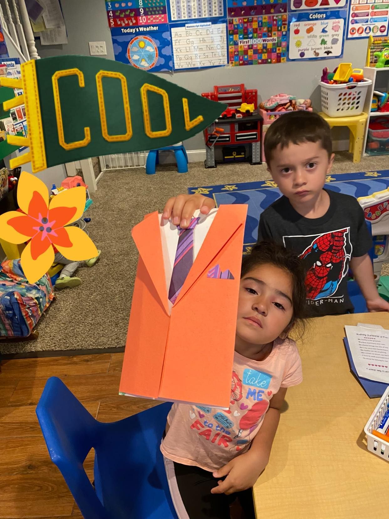 Two children in a classroom hold up colorful paper crafts and a sign reading “COOL.”