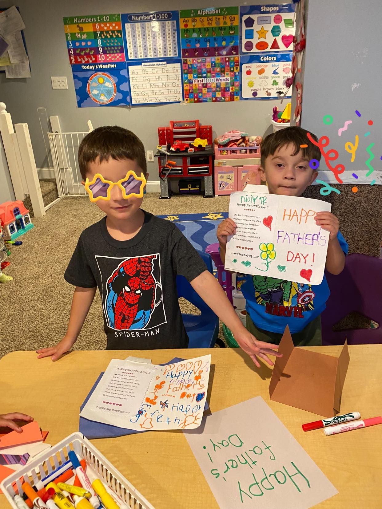 Two children in a classroom holding colorful handwritten papers at a table with markers and drawings.