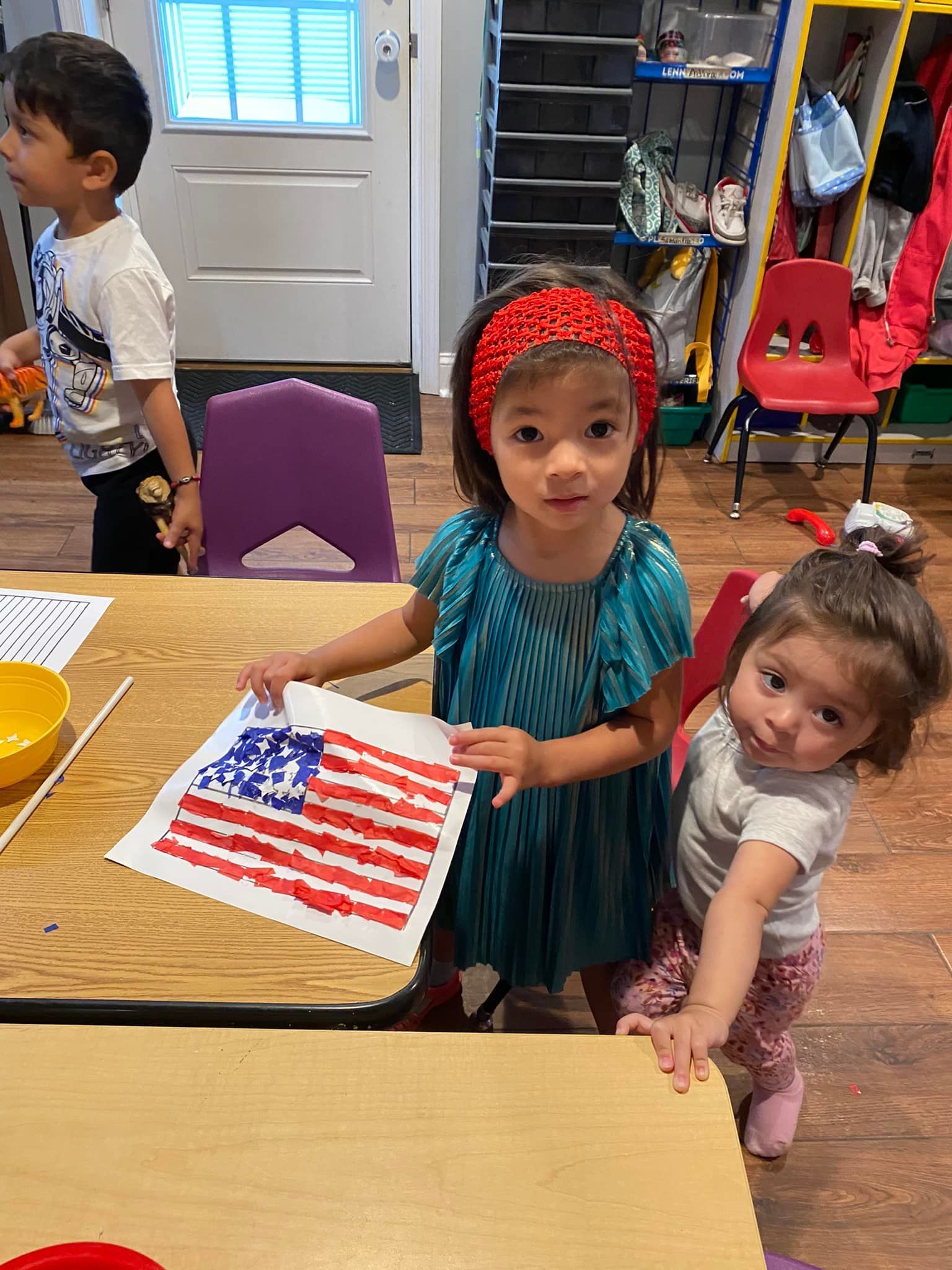 Children at a table holding a U.S. flag craft in a classroom, with chairs and toys in the background