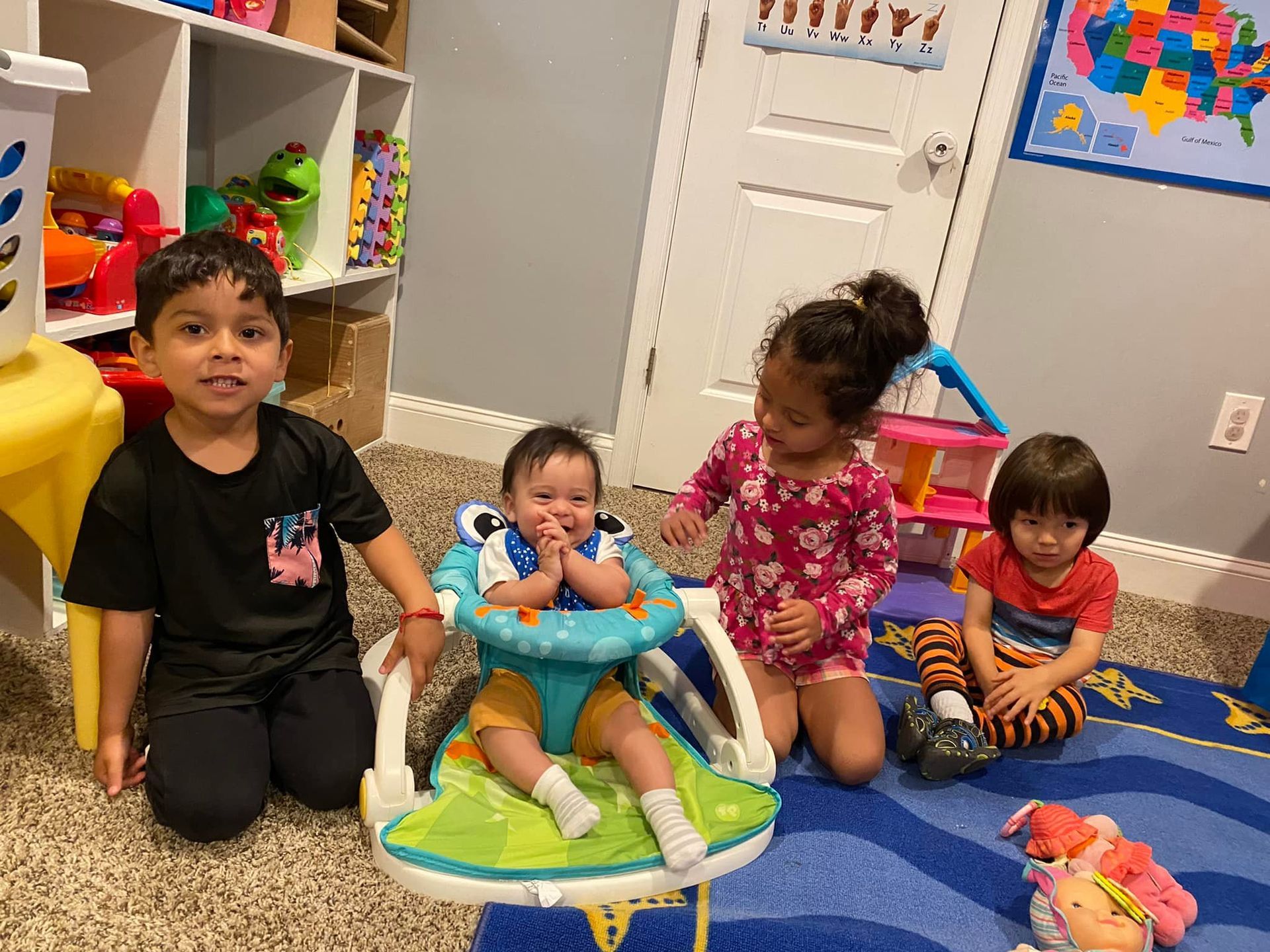 Four children playing in a playroom, sitting on colorful toys with shelves and a wall map in the background.