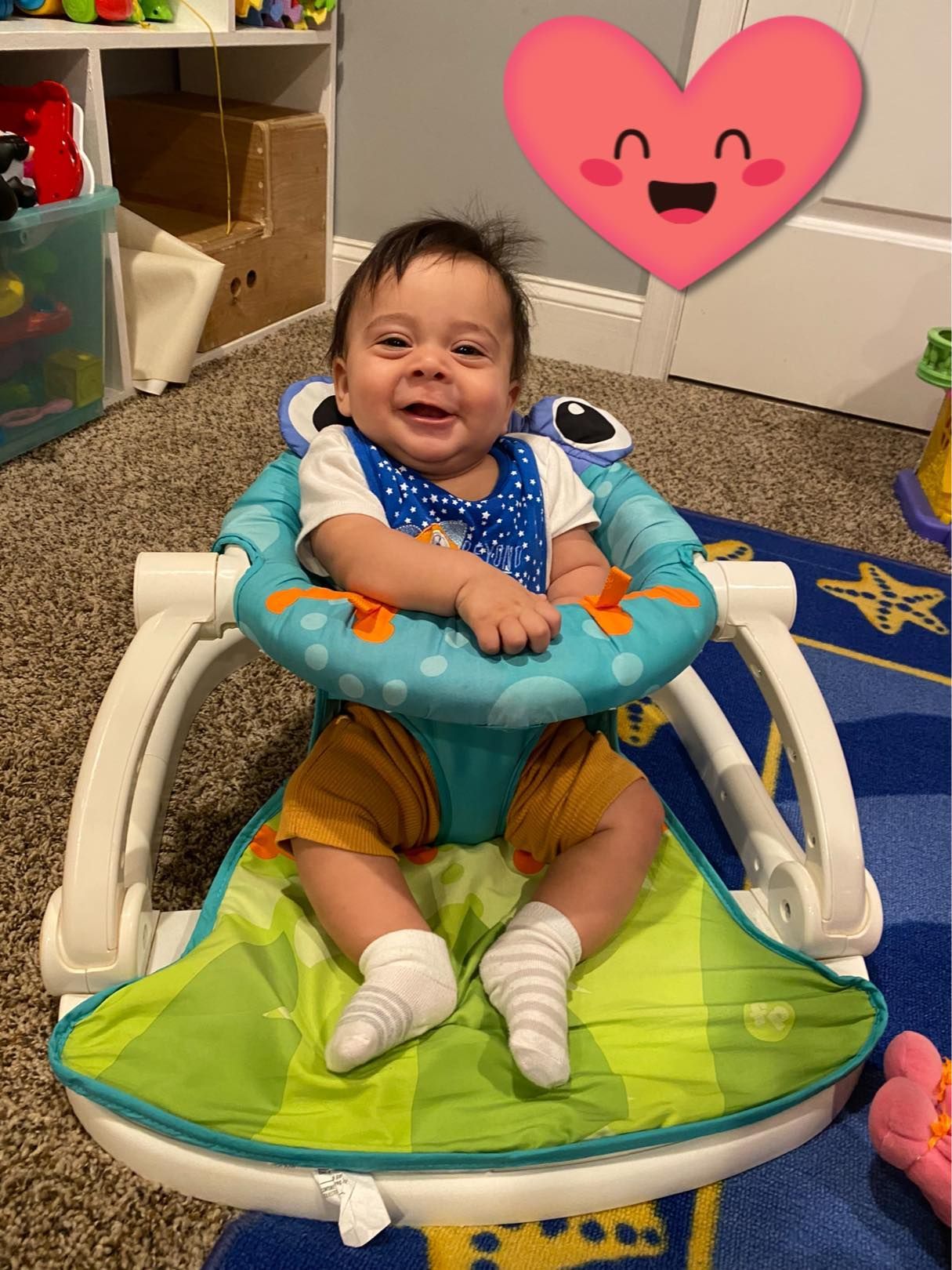 Smiling baby in a turquoise activity seat on a carpet, with a pink heart graphic above.