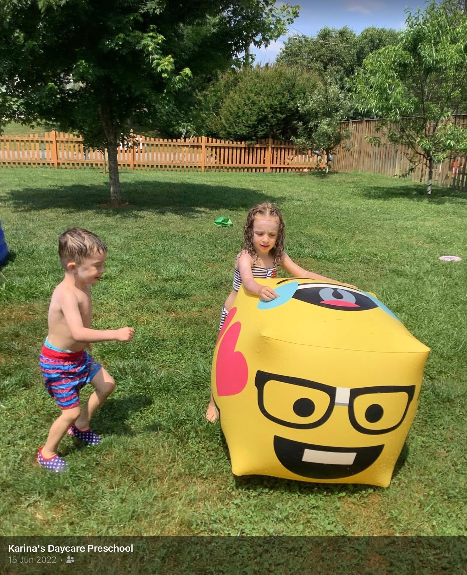 Children playing outdoors with a yellow LEGO cube toy on grass, near trees and a fence.