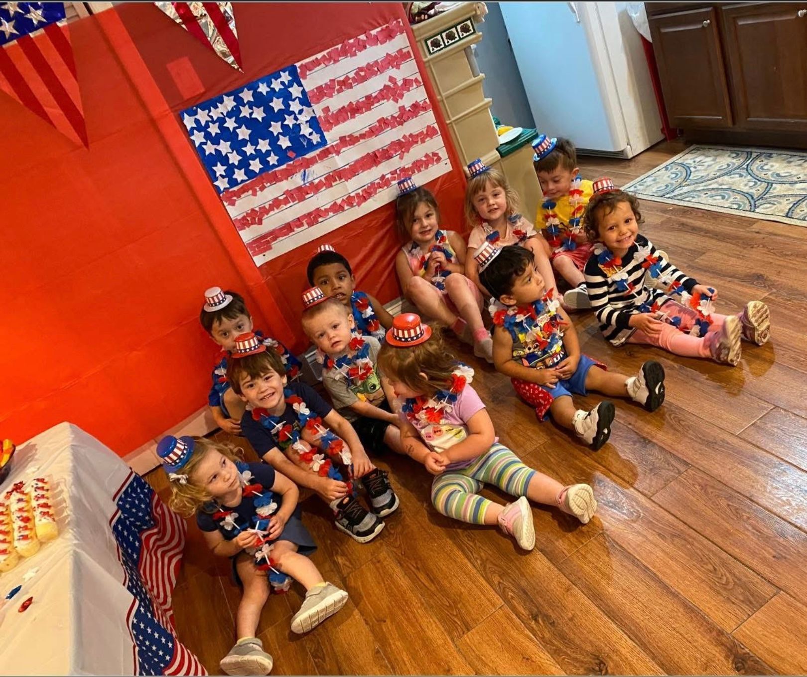 Children seated on a classroom floor beside an American flag display, wearing red, white, and blue outfits