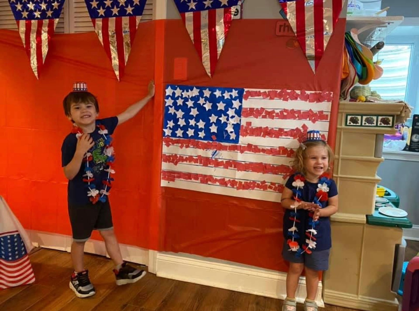 Two children posing by a wall with American flag decorations, one waving and one holding a small flag.