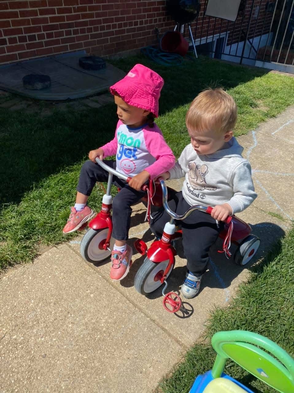 Two toddlers sit on red tricycles on a sunny sidewalk, one in a pink hat and one in a gray hoodie.