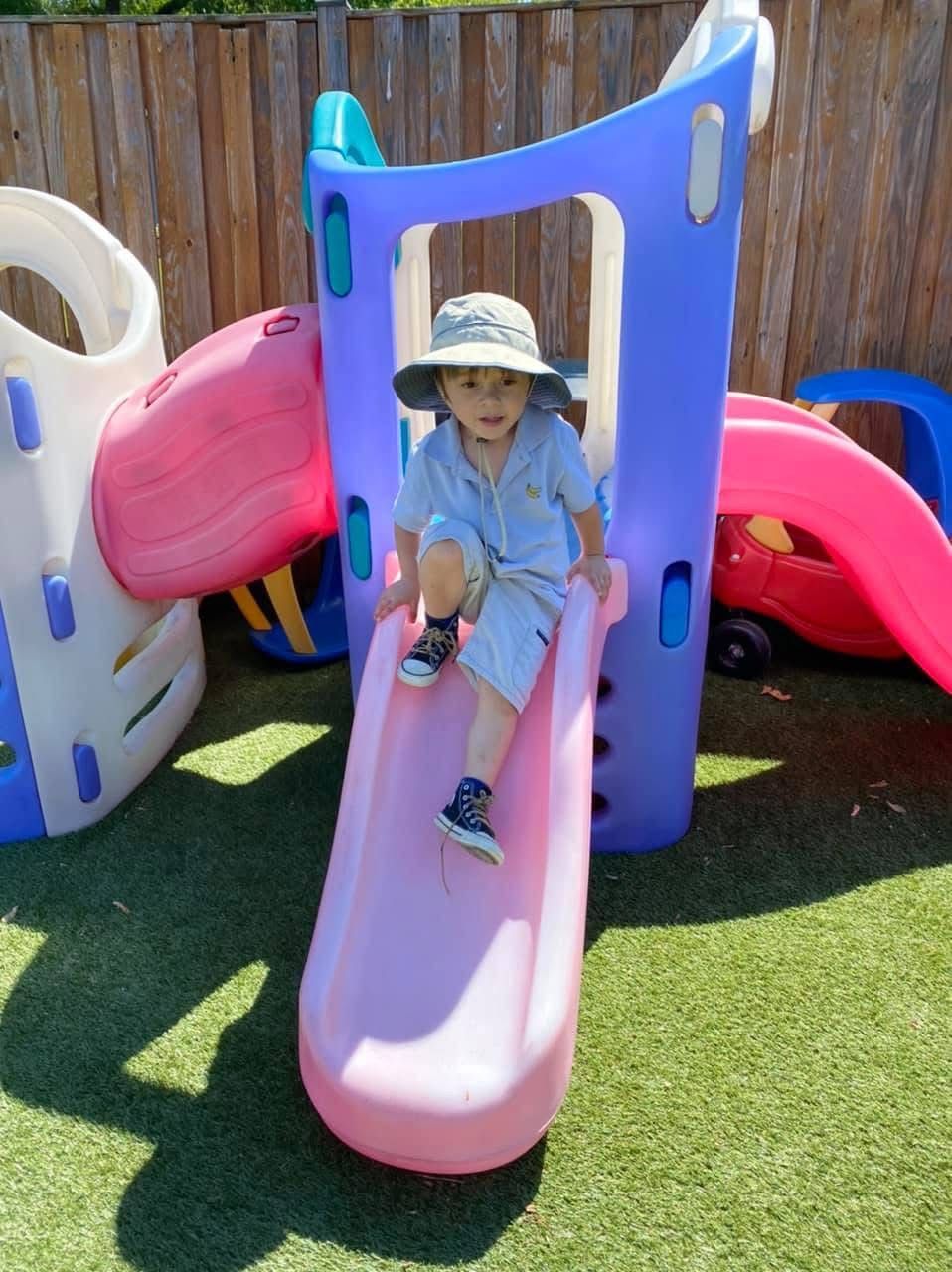 Child sitting on a pink slide in a colorful playground with artificial grass and a wooden fence.