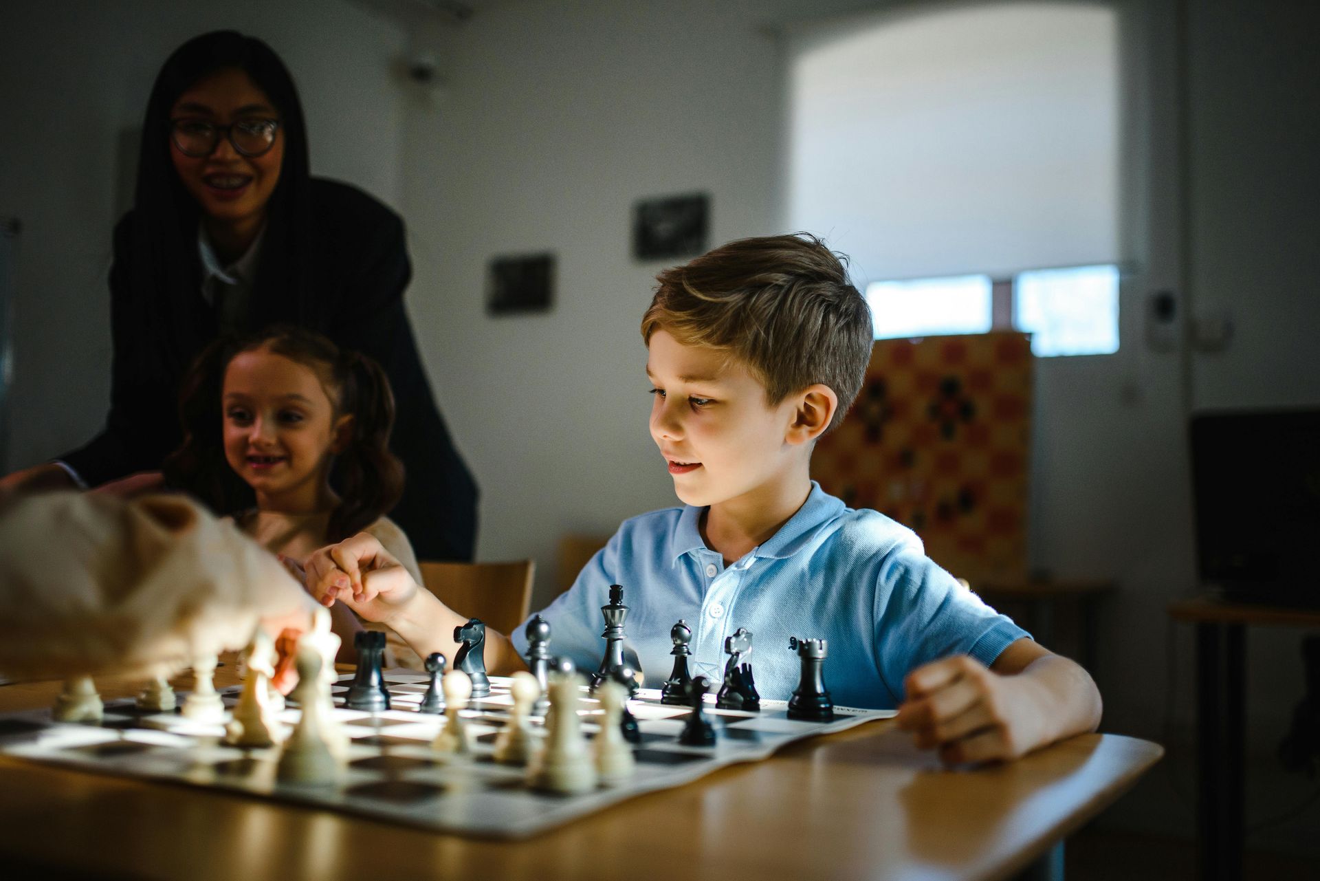 Children playing chess at a table while an adult watches nearby in a sunlit room
