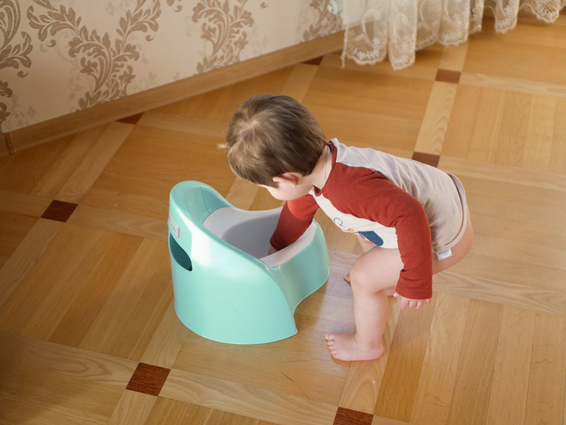 Child bending over a turquoise potty in a room with wooden floor and wallpaper