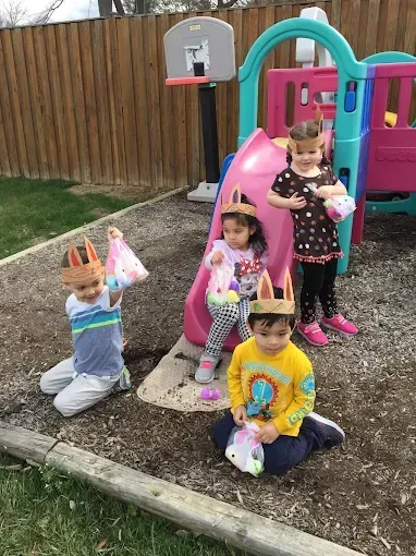 Four children in paper crowns pose by a backyard playset, holding small bags and smiling.