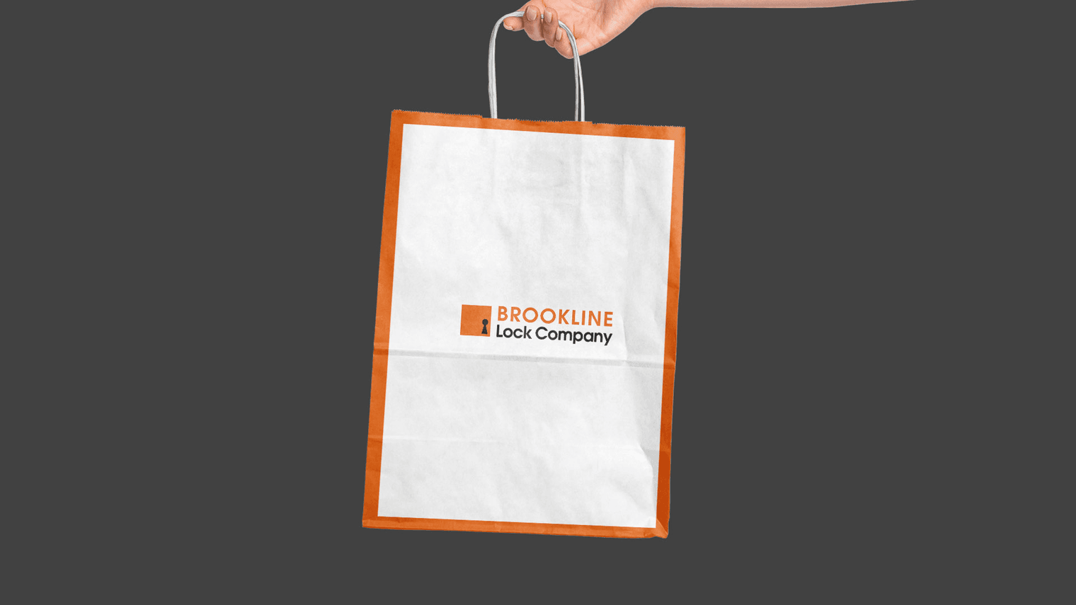 A hand holds a white paper shopping bag featuring an orange frame and the Brookline Lock Company logo