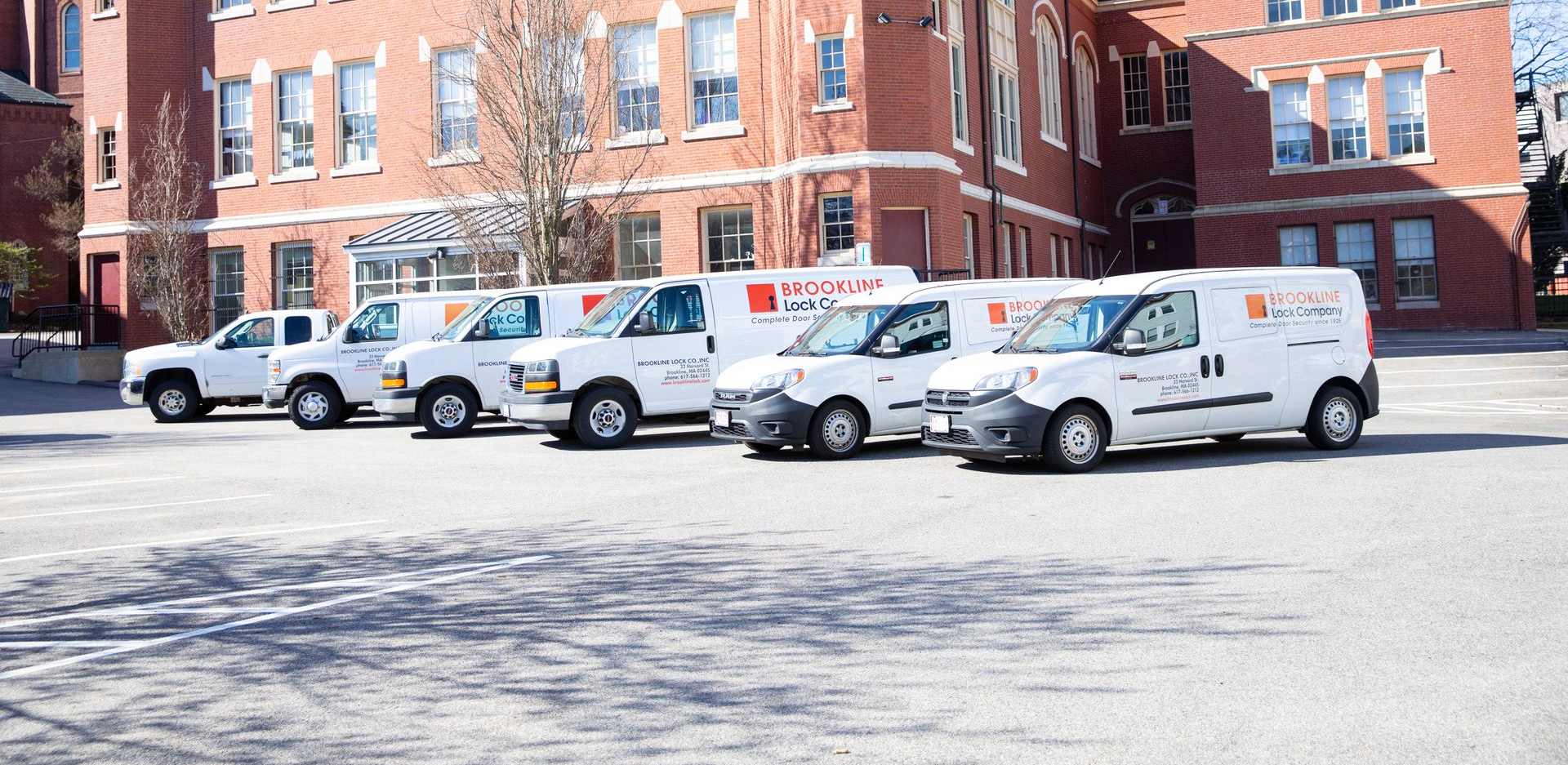 A row of white service vans parked in front of a red brick building on a sunny day.