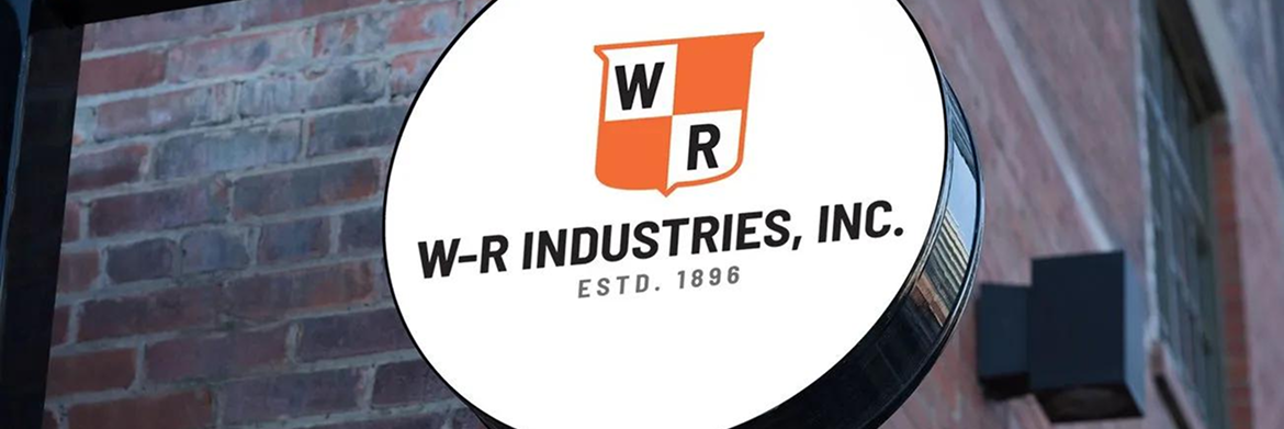 A circular sign for W-R Industries, Inc. on a brick wall, featuring an orange shield logo with a white