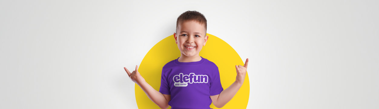 Smiling child in a purple t-shirt with a white graphic standing in front of a yellow circle, making a rock-on hand gesture.