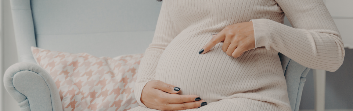 A person wearing a beige long-sleeved dress sits in a light blue chair, gently pointing to their pregnant belly.