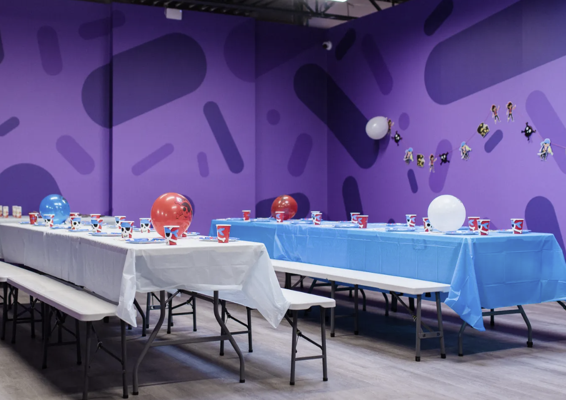 Two rows of party tables with white and blue tablecloths and balloons against a purple wall with a pattern.