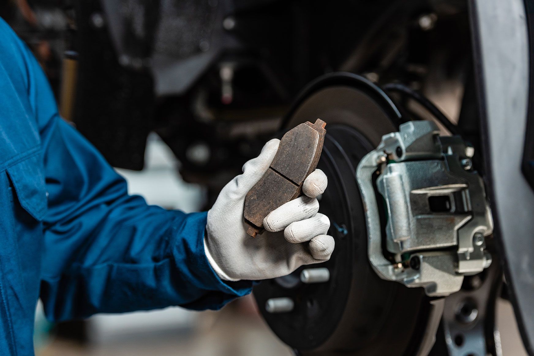 Mechanic holding worn brake pad next to brake disc assembly. Blue jumpsuit, white gloves, garage setting.