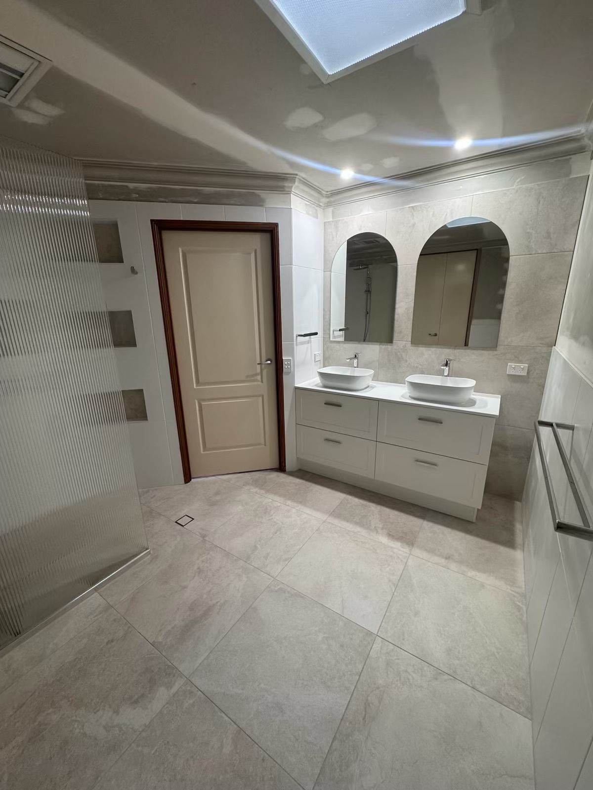 Bathroom with white vanity, arched mirrors, glass shower, and neutral tile.