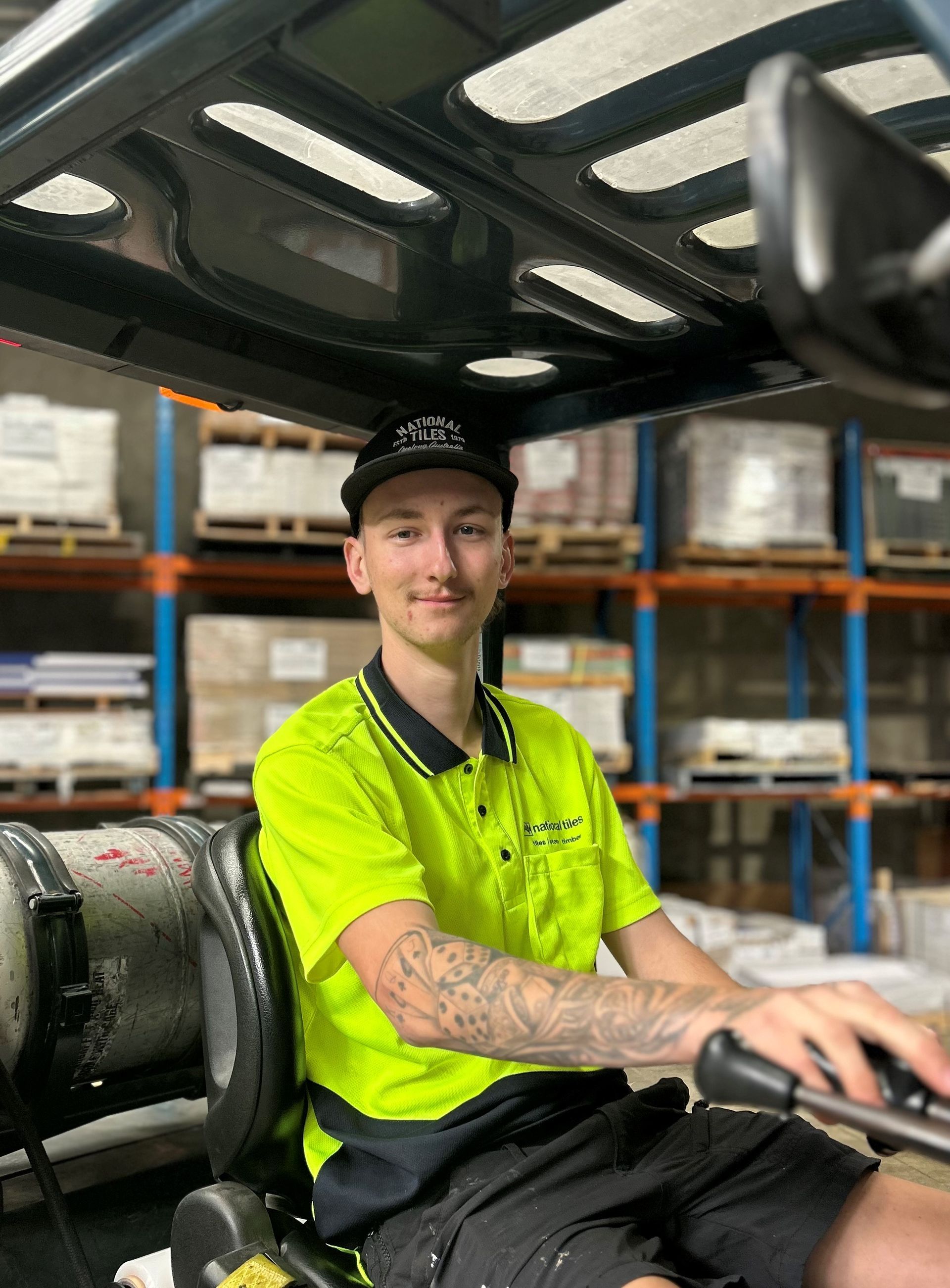 Young man driving a forklift in a warehouse, wearing a neon green shirt and a black cap.