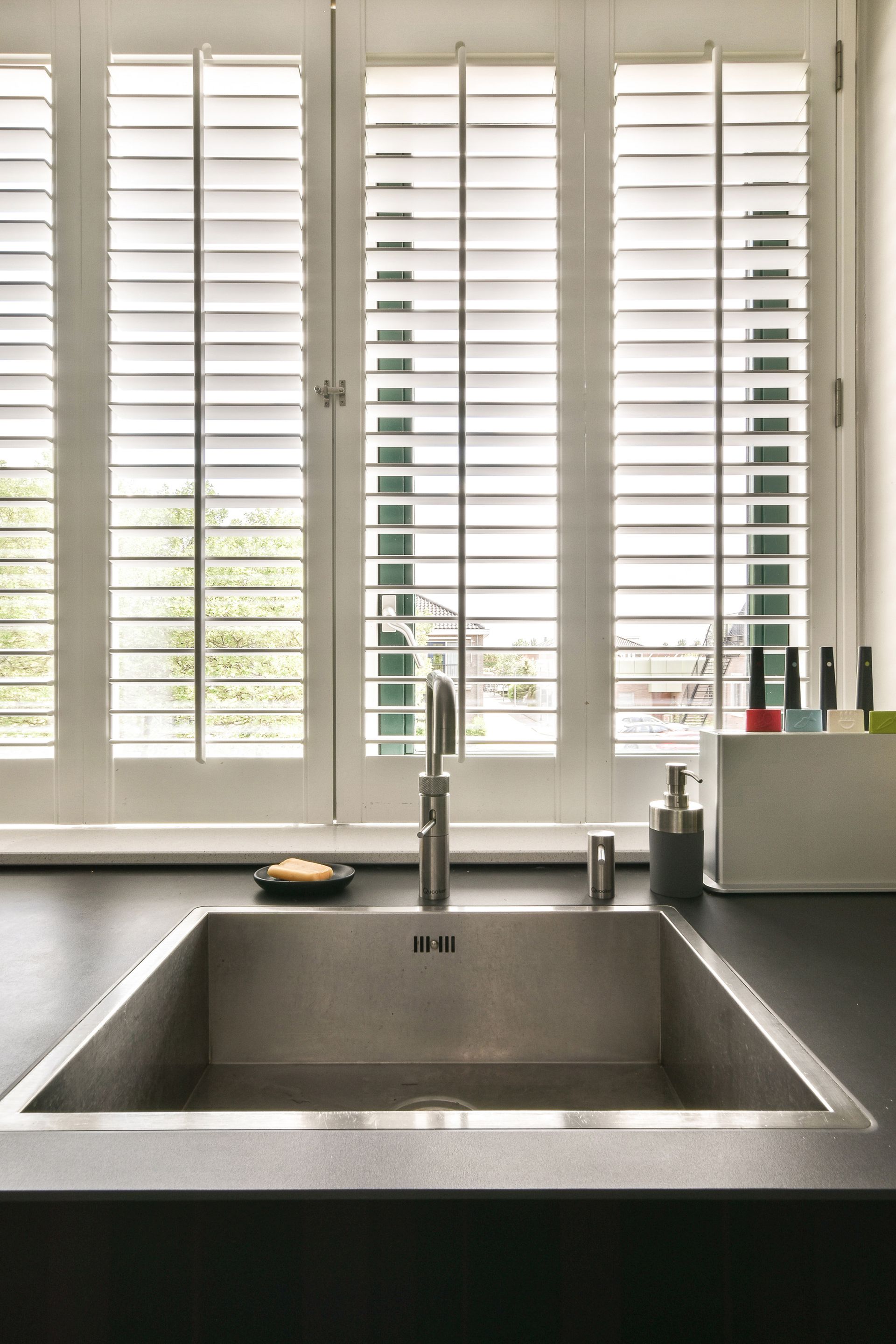 Stainless steel kitchen sink under white shuttered window.