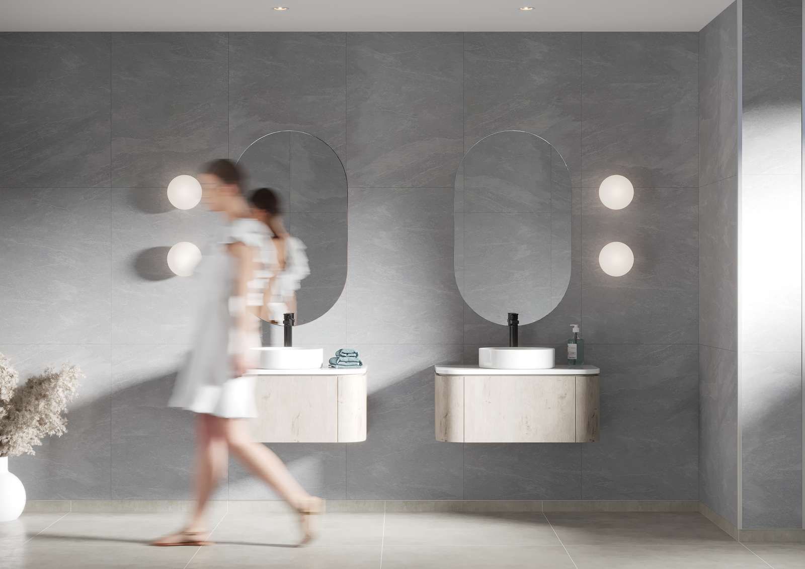 Modern bathroom with gray walls, two sinks, oval mirrors, and a person walking by.