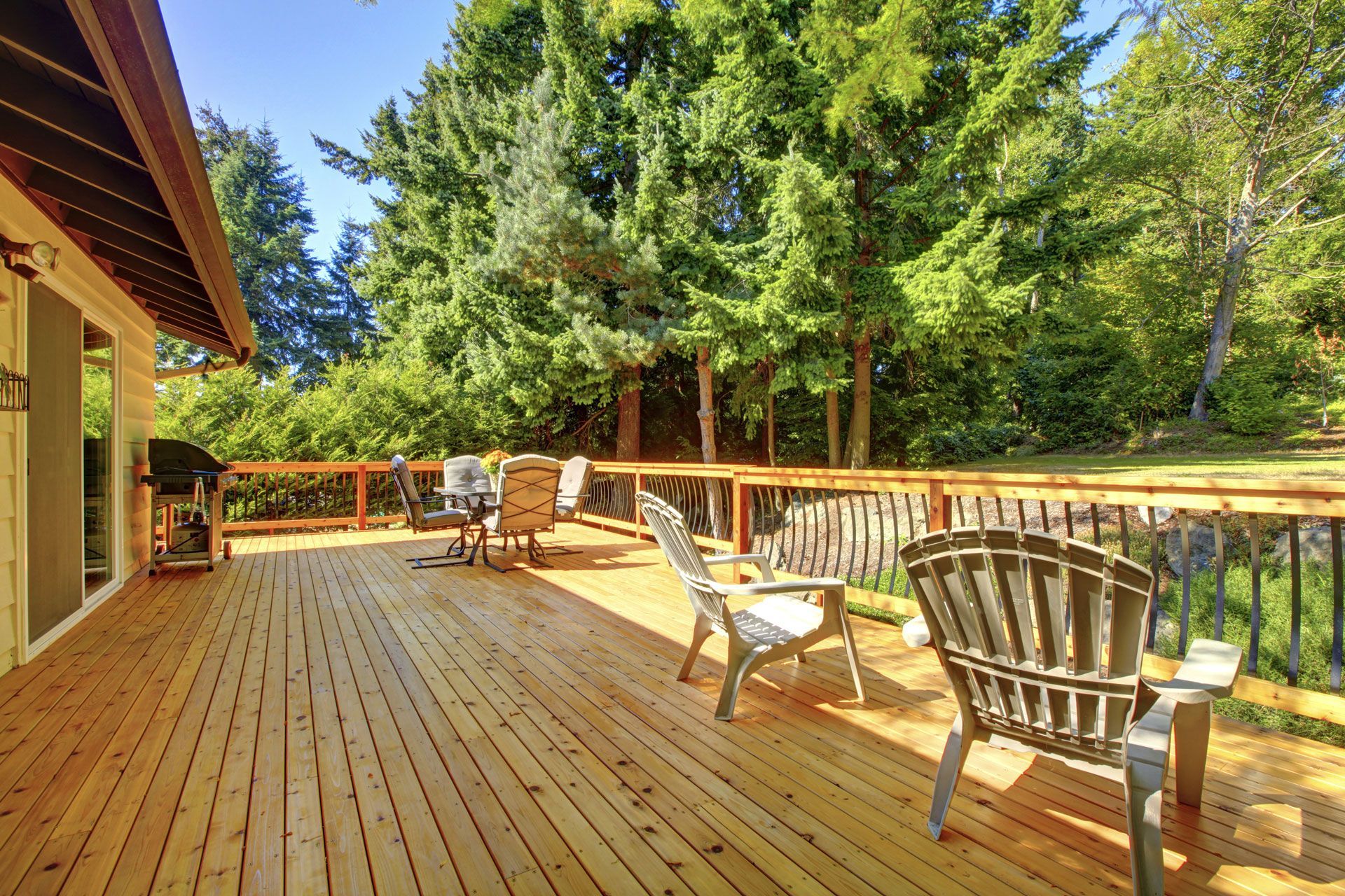Wooden deck with chairs, table, and grill. Lush green trees in background. Sunny day.