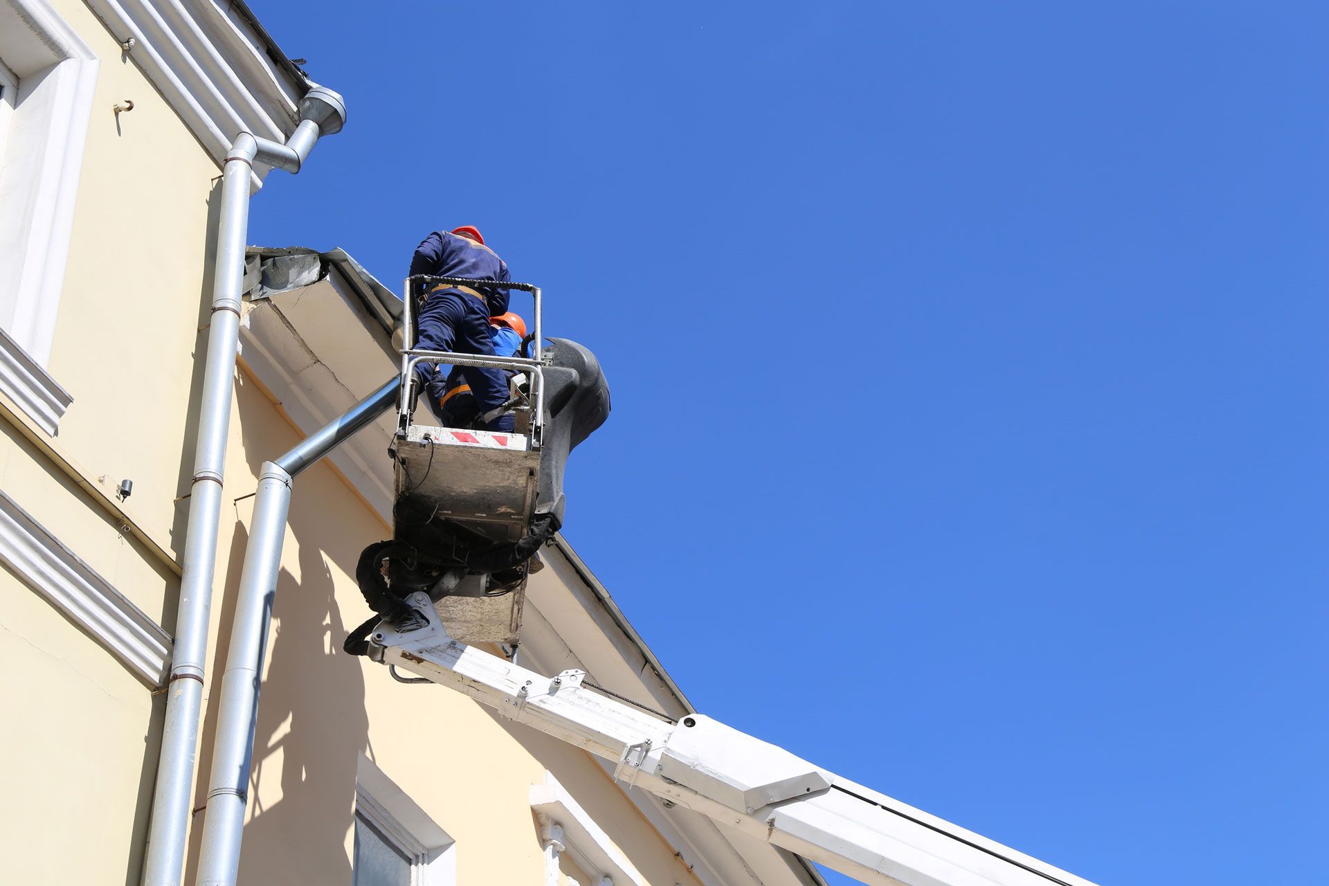 Two workers in a lift basket repairing the roof of a yellow building under a blue sky.