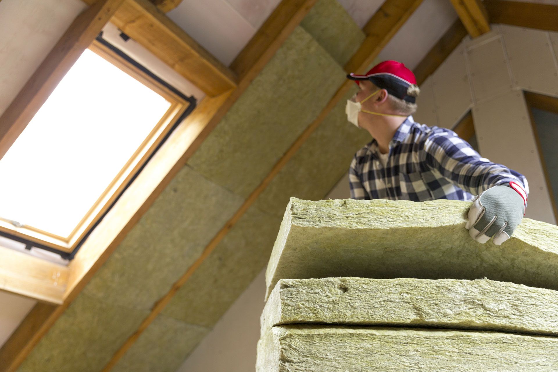Man installing insulation in an attic, wearing a respirator, gloves, and a cap.