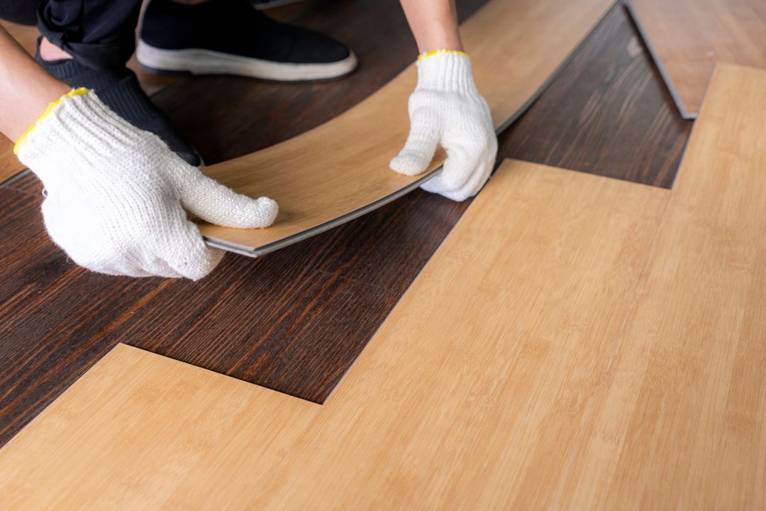 Hands in gloves installing light-colored vinyl flooring over a darker section; close-up.