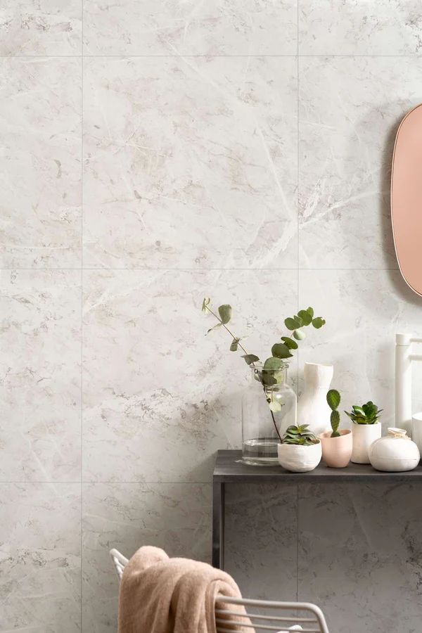 Bathroom wall with white marble-look tiles, gray shelf holding plants and vase, pink mirror, and a chair with a blanket.