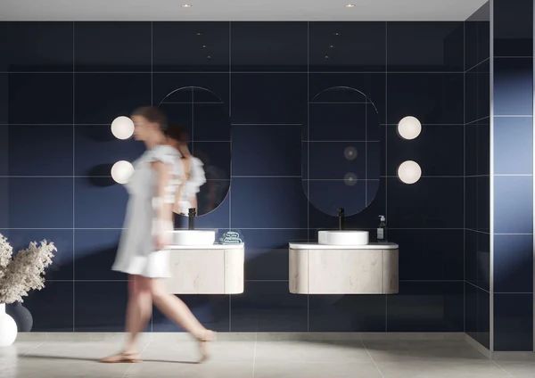 Woman walking in a modern navy blue bathroom with floating wooden vanities and round mirrors.