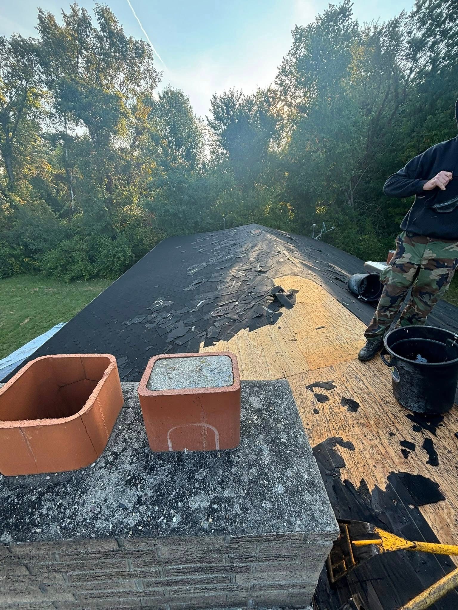 A person near a chimney on a roof with brick chimney caps and debris. Green trees and sky in the background.