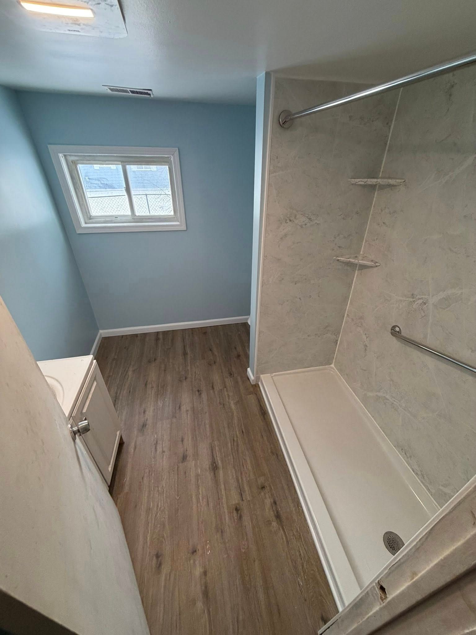 Bathroom with light blue walls, gray tile shower, wood-look floor, and small window.