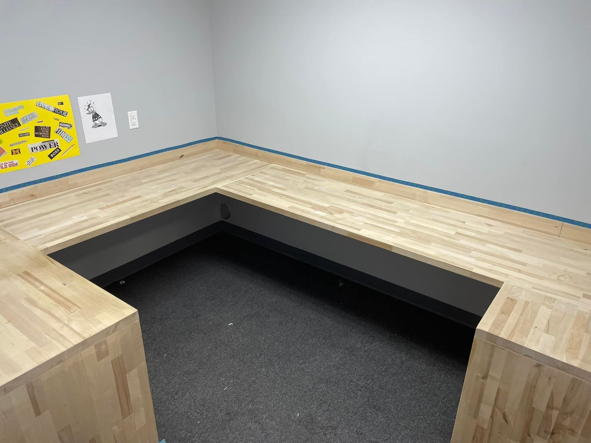 L-shaped wooden desk in a room with gray walls and a dark floor. A cutout area is under the desk.