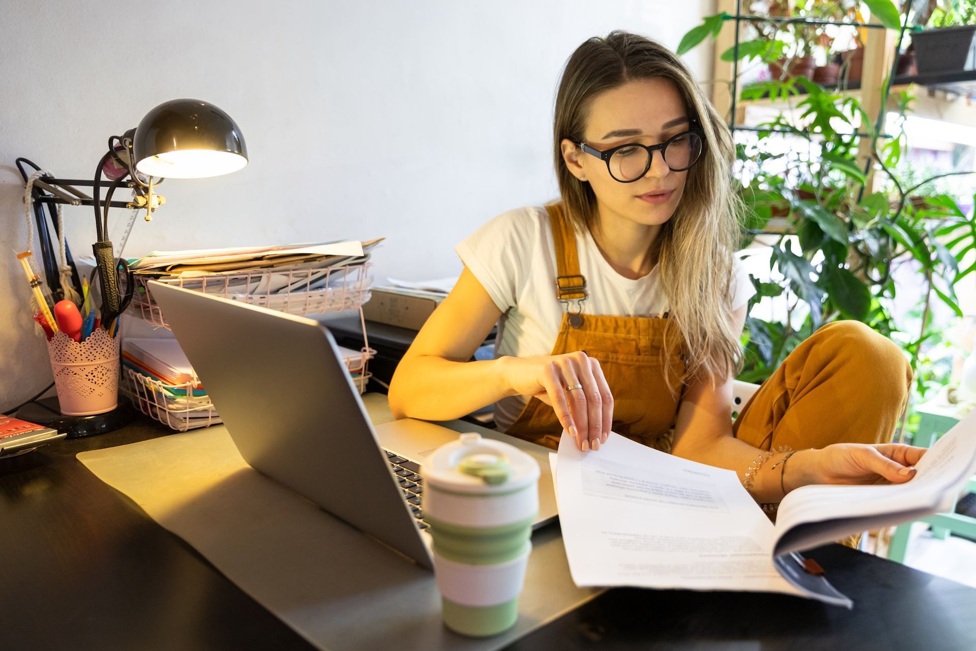 Person studying at a desk with a laptop, open book, and coffee beside a lamp in a bright room