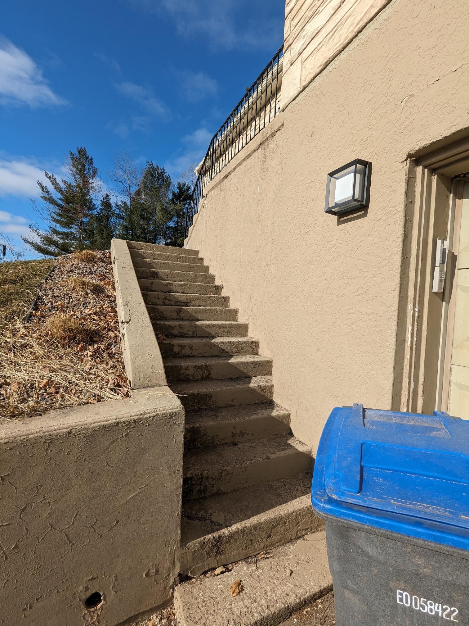 A blue trash can is next to a set of stairs leading up to a building.