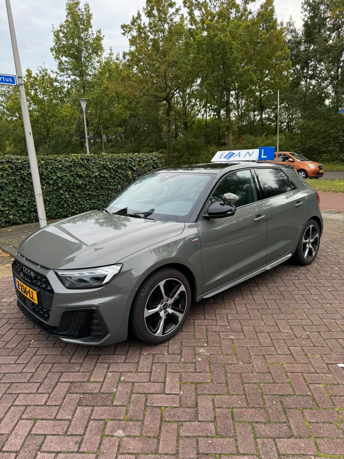 Een grijze hatchback staat geparkeerd op een bakstenen oprit naast groen, met een dakbord en een gele kentekenplaat.