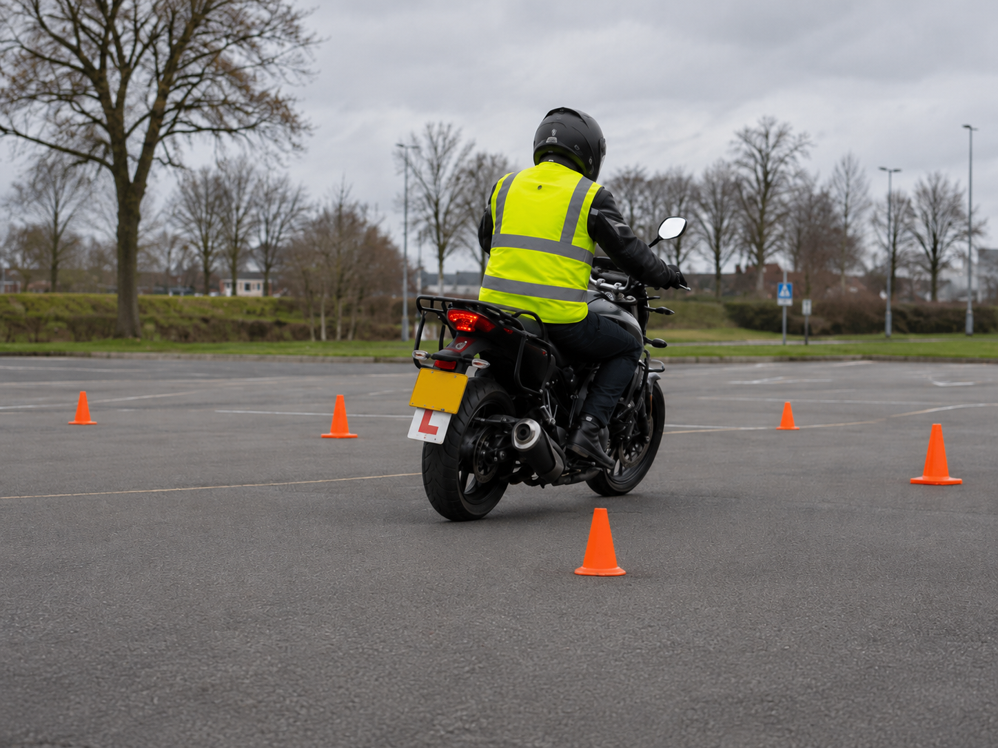 Een motorrijder in een geel hesje rijdt tussen oranje kegels door op een parkeerplaats op een bewolkte dag.