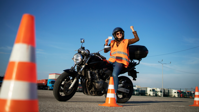 Motorrijder in oranje veiligheidsvest poseert naast een zwarte motor tussen verkeerskegels op een zonnig trainingsparcours.