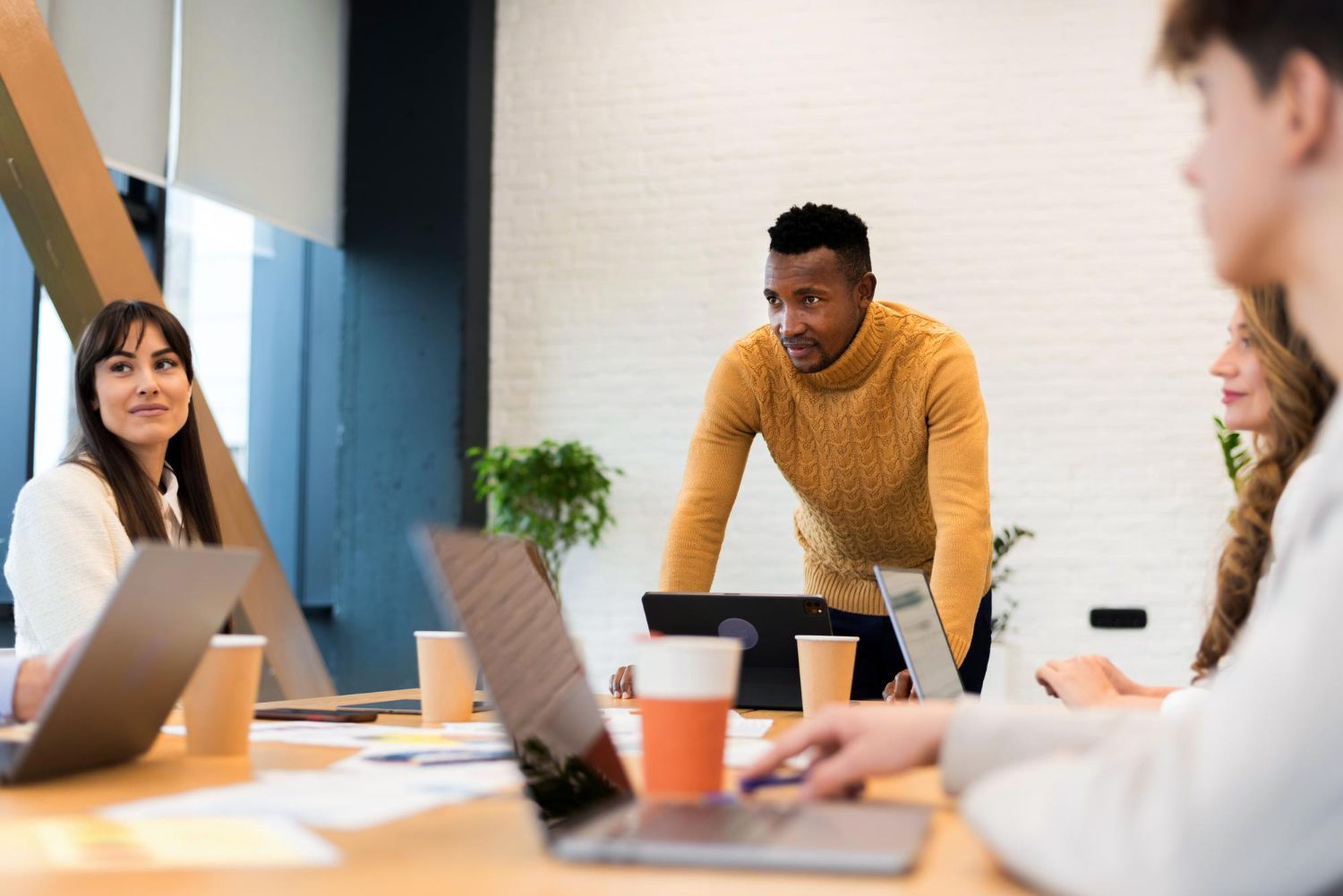 Coworkers in a meeting around laptops in a modern office, with one person standing and presenting.