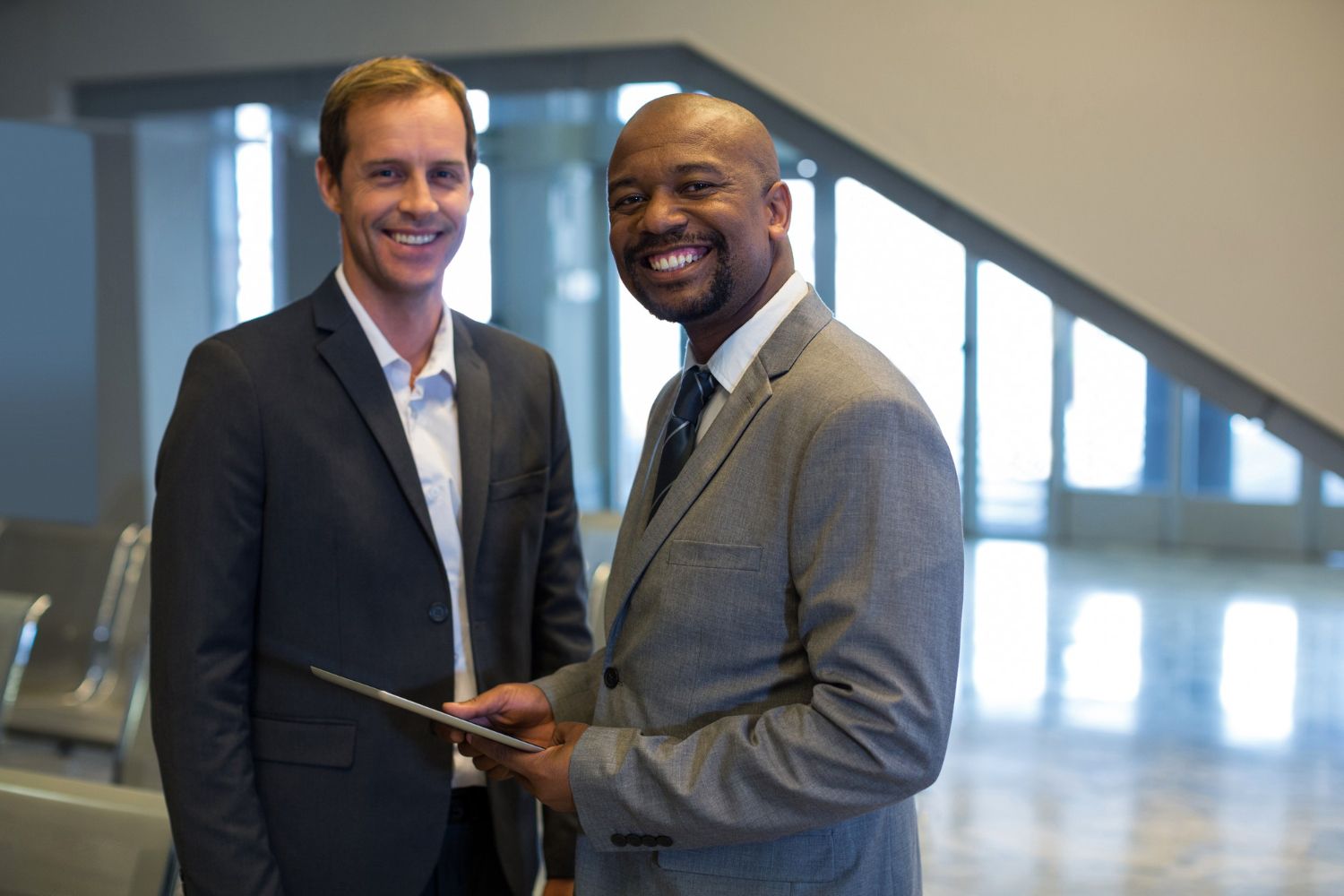 Two men in business suits smiling in a bright office lobby, holding a tablet