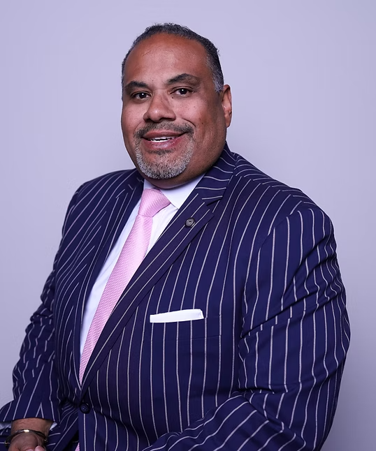 Portrait of a man in a navy pinstripe suit with a pink tie, smiling against a light purple background