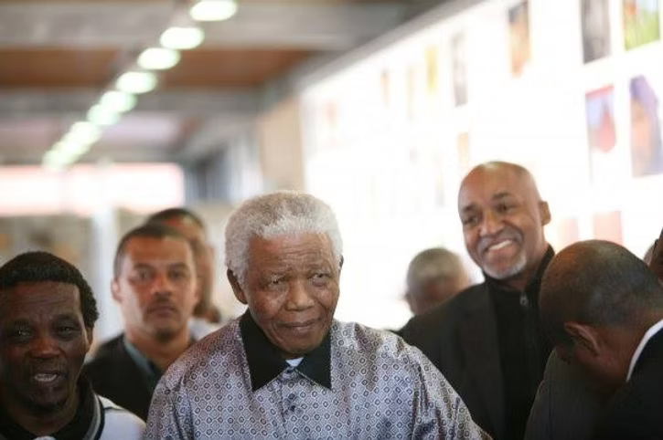 Nelson Mandela smiles while standing among a small group of people indoors.