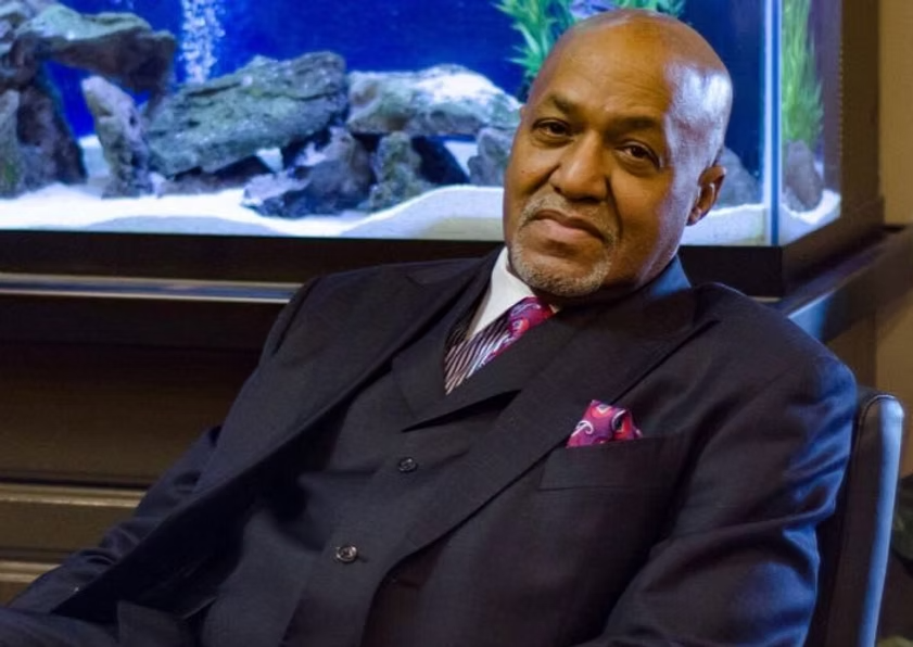 Man seated in a chair in front of an aquarium, wearing a dark suit and pink tie, looking at the camera