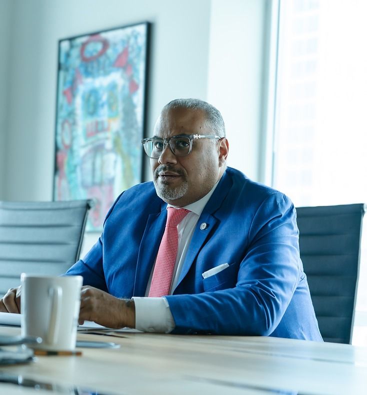 Man in a blue suit and pink tie seated at a conference table in a bright office