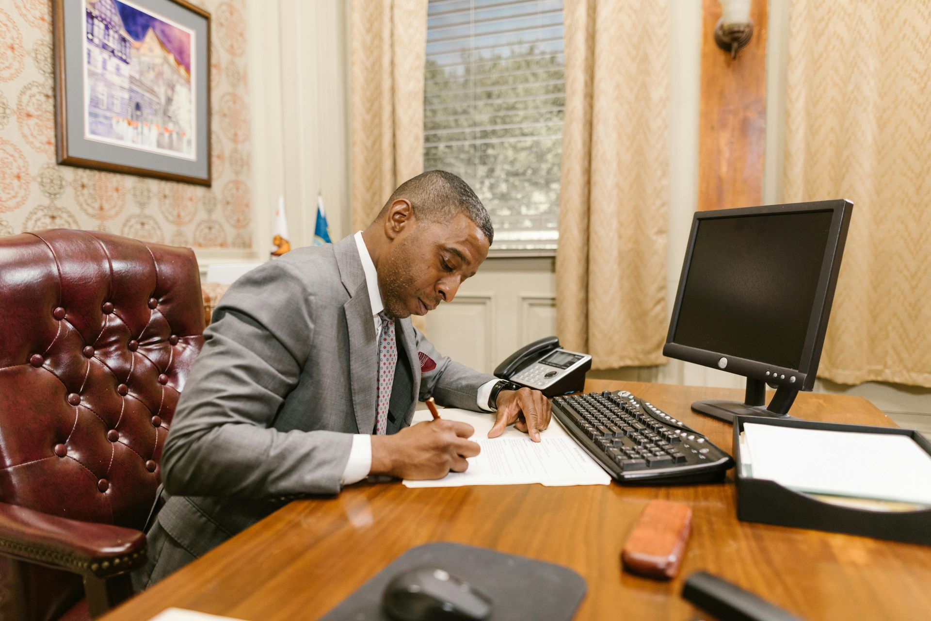 Man writing at a desk in an office beside a computer monitor and phone.