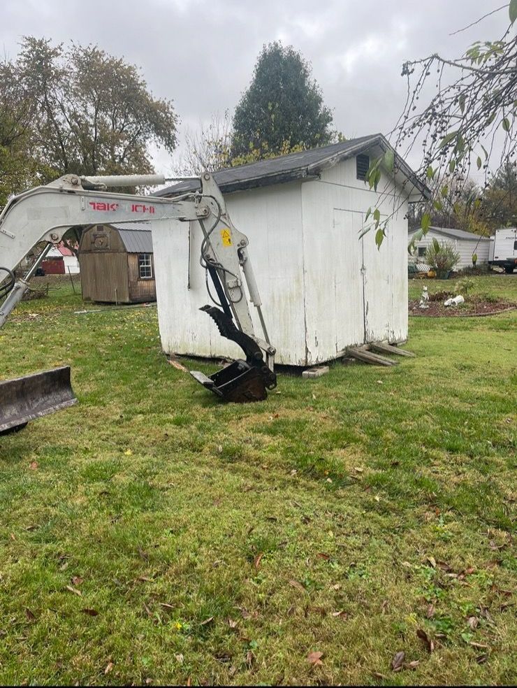 An excavator is sitting in the grass in front of a white shed.
