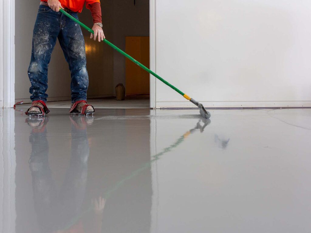 Person in paint-splattered jeans smoothing gray epoxy flooring with a long-handled tool in a room.
