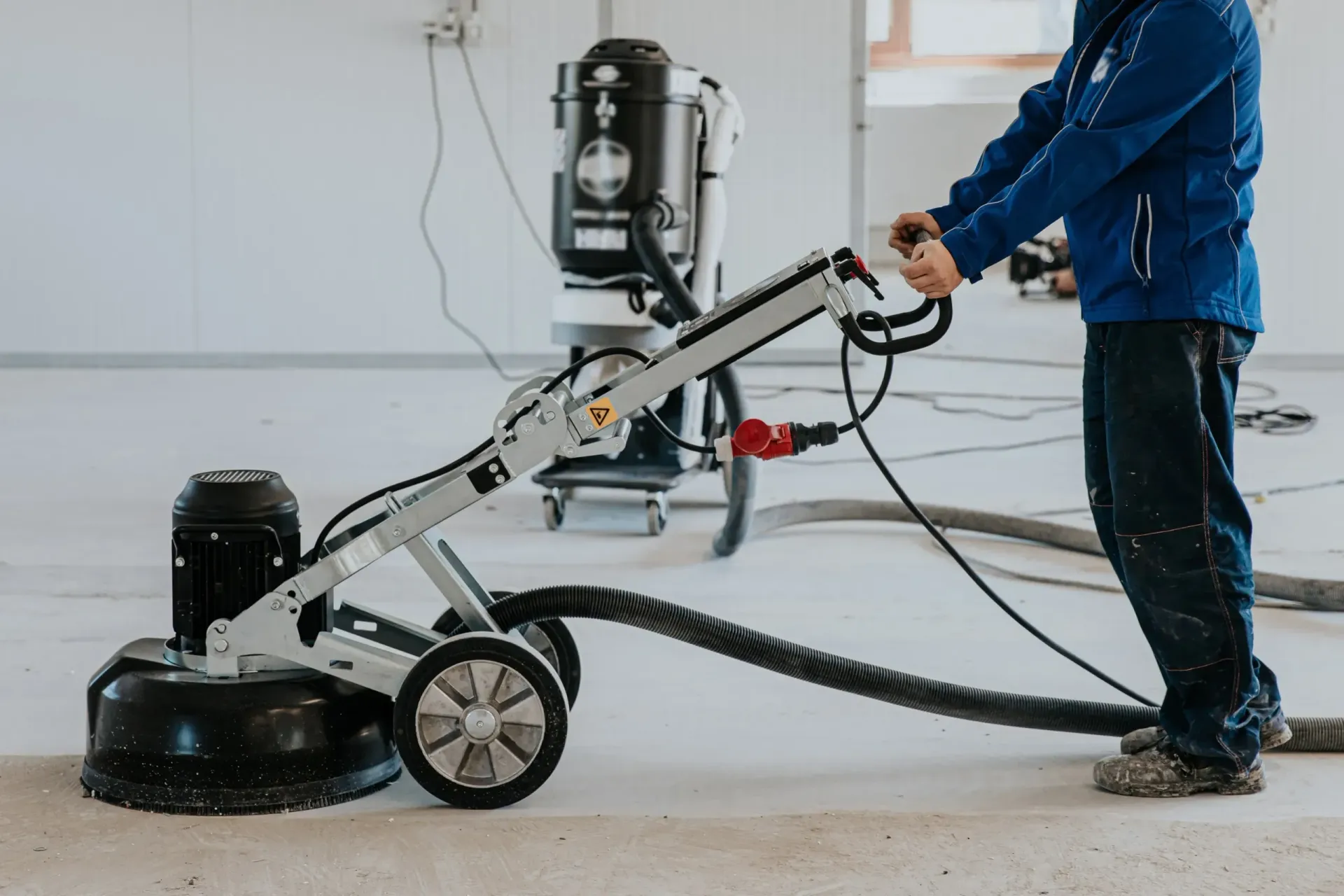 Person using a concrete floor grinder in a light-colored interior. The machine has a black dust collection system.