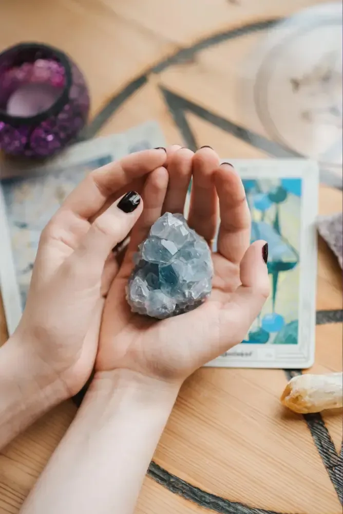 Hands holding a blue-green crystal over a wooden table with tarot cards and candles nearby
