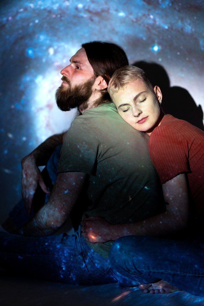Two people embracing amid a starry blue cosmic background, one resting asleep on the other's shoulder